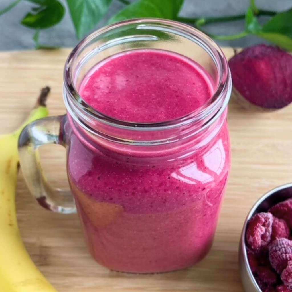 Bright pink raspberry beet smoothie in a mason jar on a wooden board, with frozen raspberries, a banana, and a whole beet nearby