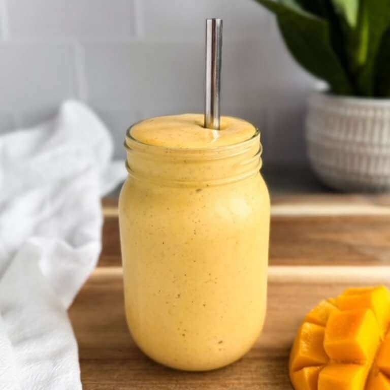 Creamy mango turmeric smoothie in a glass jar with a metal straw on a wooden board with fresh mango and white cloth beside it