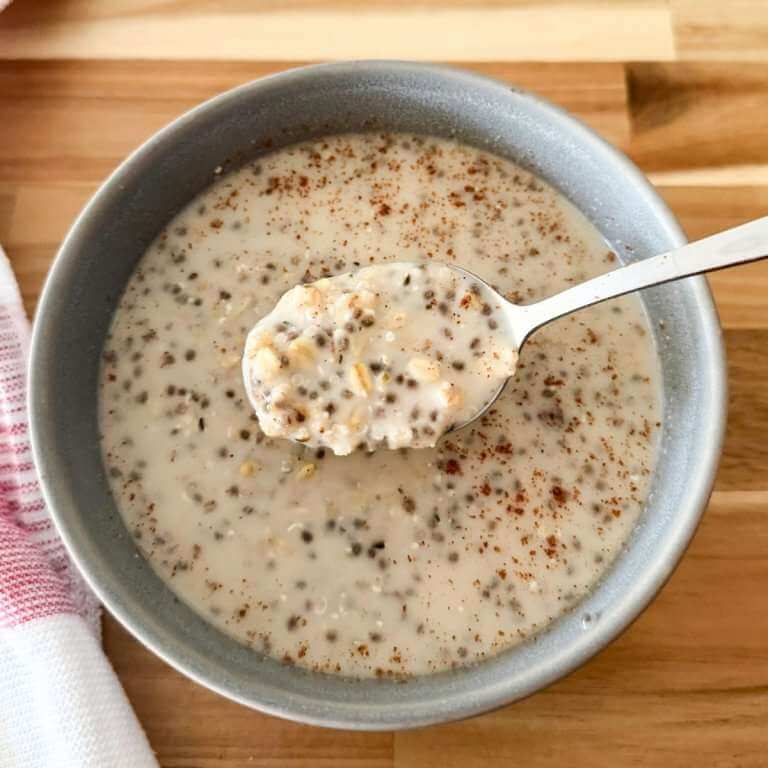 Spoon lifting creamy oatmeal with chia seeds and rolled oats from a bowl of oatmeal topped with cinnamon on a wooden board