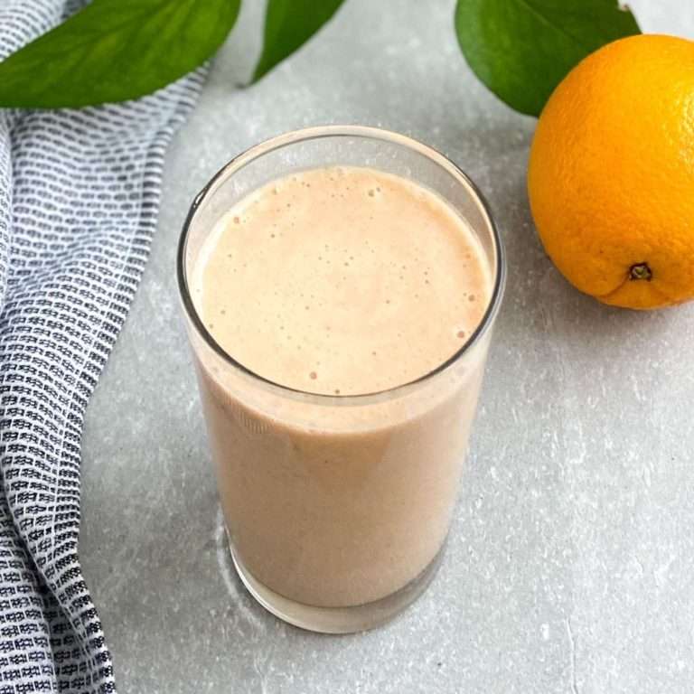 Banana orange smoothie in a glass cup with a orange, tea towel and plant leaves surrounding the smoothie.