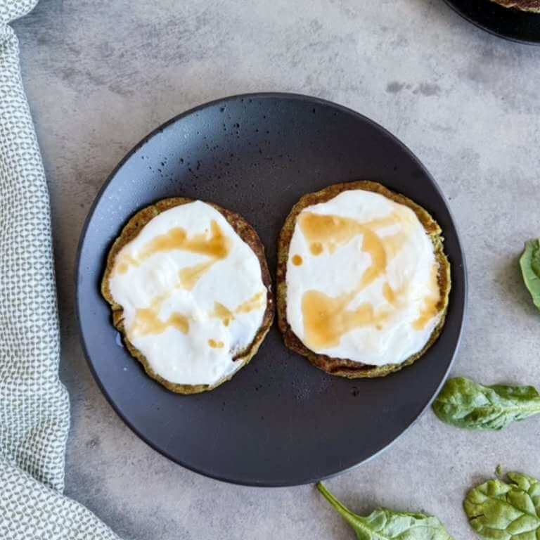 Two spinach pancakes topped with yogurt and a drizzle on a dark plate with spinach leaves on a light countertop