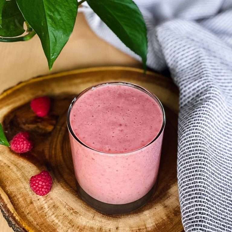 raspberry banana smoothie in a glass cup on a wood chopping board with a tea towel and plant on the side
