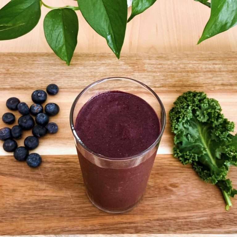 kale and blueberry smoothie in a glass cup on a wood chopping board with fresh blueberries and a kale leaf on the side of the glass