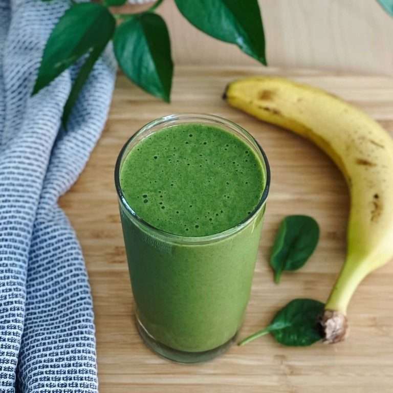 banana pineapple spinach smoothie on a wood table with a tea towel, plant leaves, spinach leaves and banana on the side of the smoothie cup