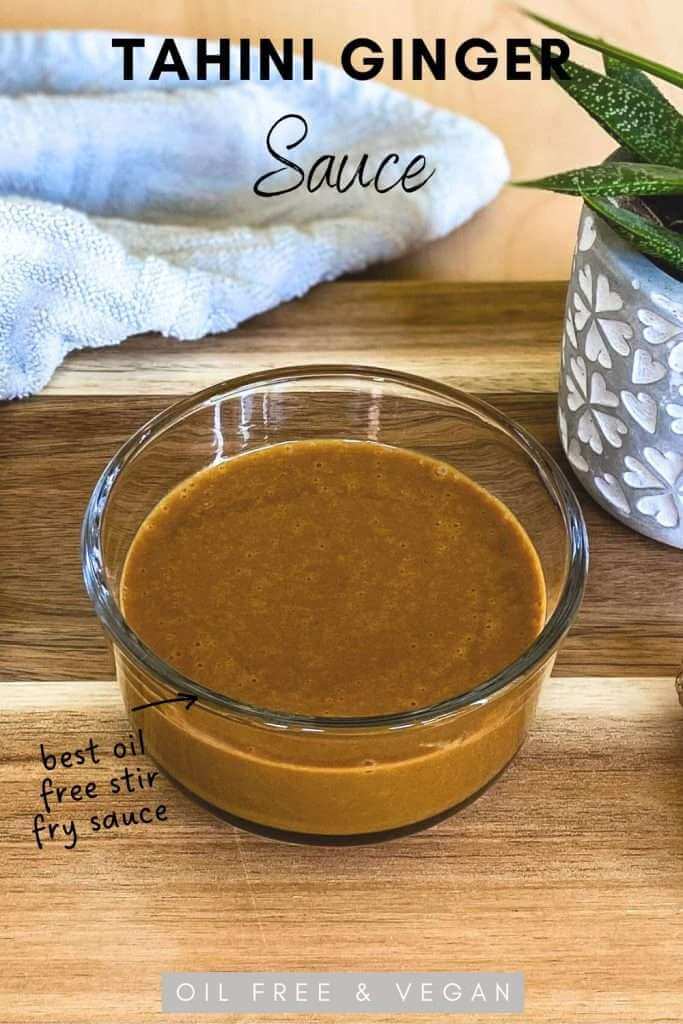 Oil Free and vegan tahini ginger sauce in a glass ramakin on a wood table with a small plant and tea towel in the background.