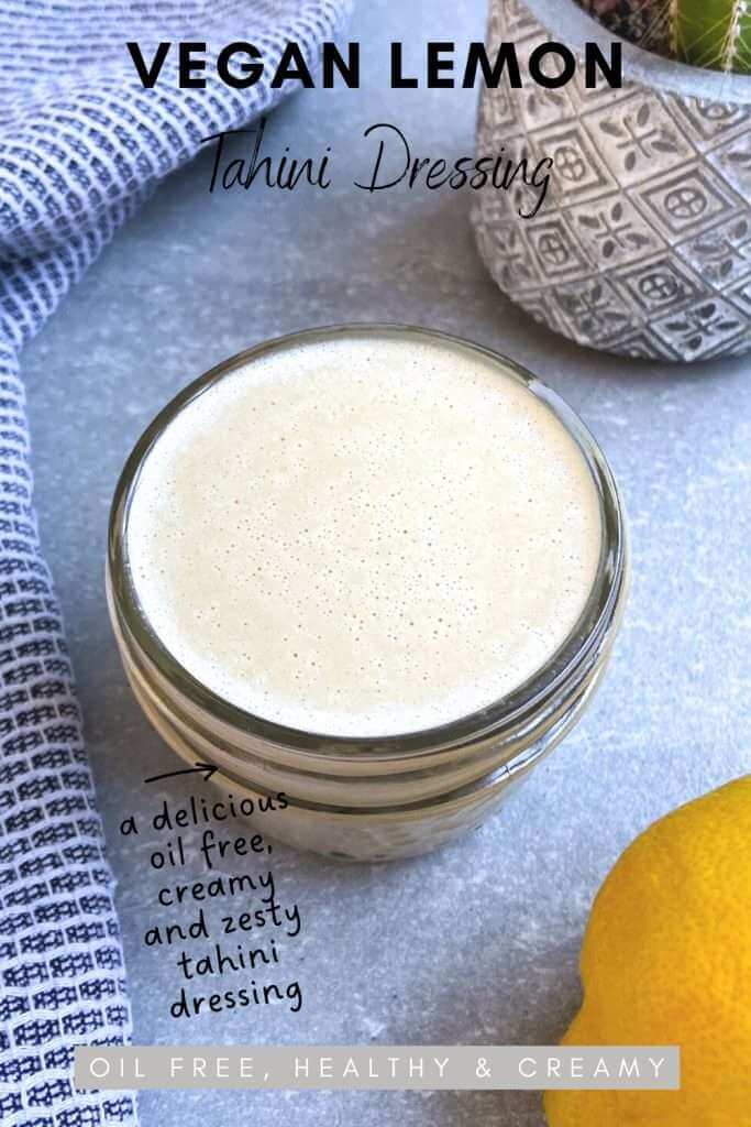 Vegan lemon tahini dressing in a glass mason jar on a concrete backdrop with a lemon, tea towel and cactus plant beside the oil free dressing.