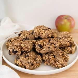 A plate of homemade oatmeal cookies with raisins and apple pieces, with a red and green apple in the background.
