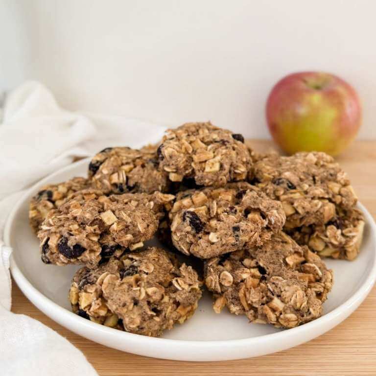 A plate of homemade oatmeal cookies with raisins and apple pieces, with a red and green apple in the background.