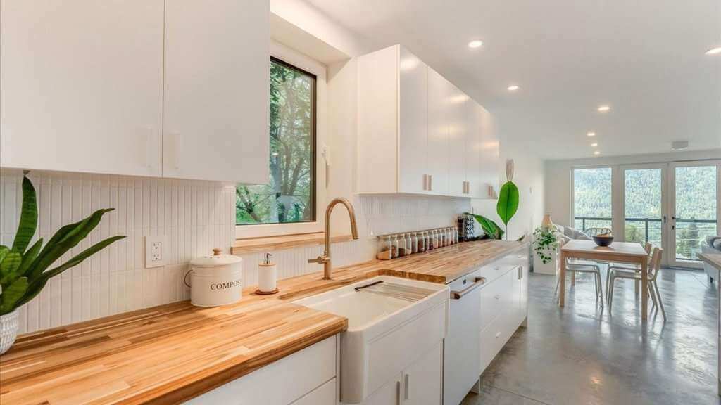 Bright modern white kitchen with wood countertops, farmhouse sink, houseplants, dining area, and mountain views through large windows.