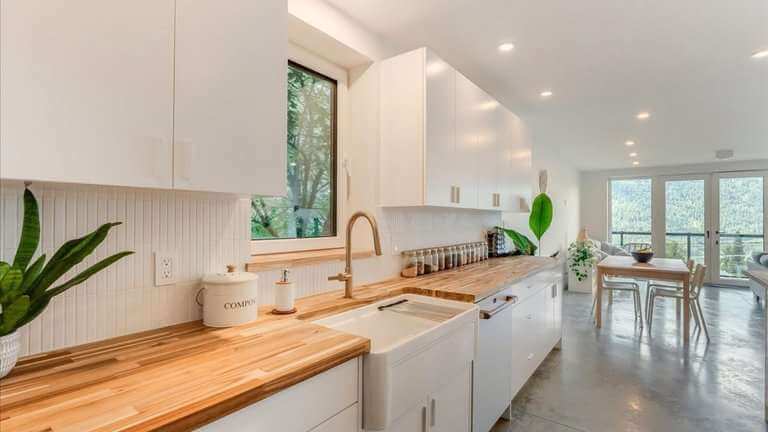 Bright modern white kitchen with wood countertops, farmhouse sink, houseplants, dining area, and mountain views through large windows.