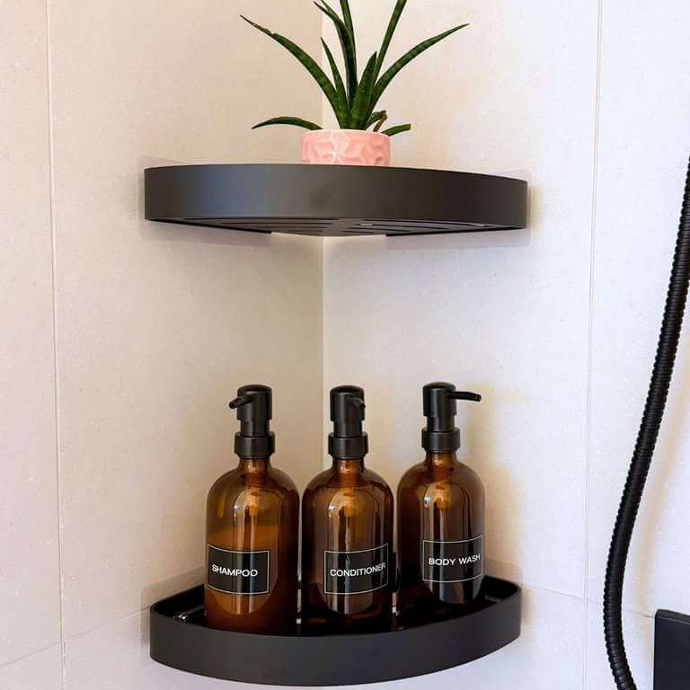 Two matte black corner shower shelves with a small potted plant and three amber glass bottles labeled shampoo, conditioner, and body wash.